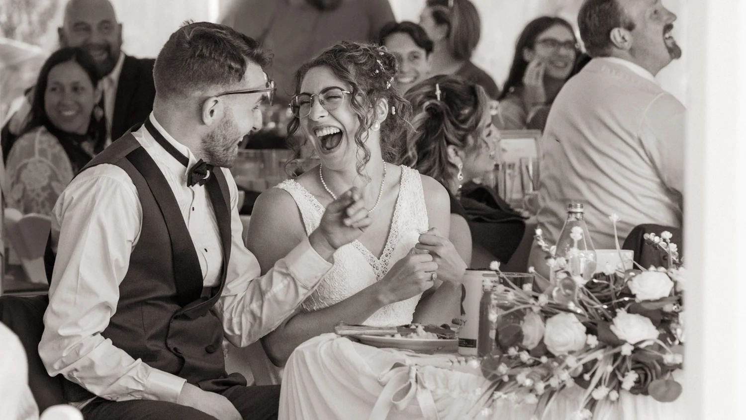Des mariés attablés qui rient, leurs invités derrière eux